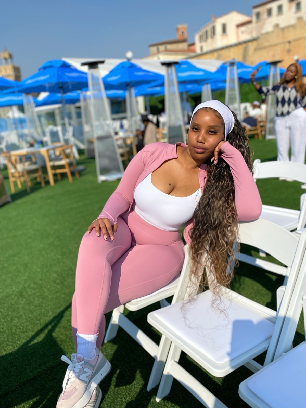 Oratile, seated among white chairs at an outdoor event, dressed in soft pink.